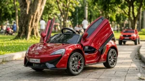 A photorealistic 4K close-up of a sleek, deep candy-apple red electric ride-on sports car for toddlers, with both butterfly-style doors wide open, parked on a stone pathway in a sunny park.