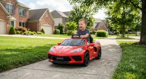 Toddler driving a mini Chevrolet Corvette ride-on car along a sunny neighborhood sidewalk.