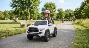 Young child wearing a safety helmet driving a white Toyota Tacoma ride-on toy truck on a paved park trail