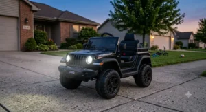 Black 12V Jeep Wrangler ride-on toy car with bright LED headlights turned on at dusk