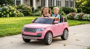 Two children smiling while riding together in a pink 24V Range Rover ride-on car in a sunny backyard.